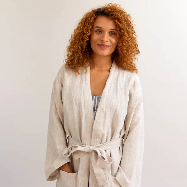 Curly haired woman smiling and wearing the Piglet in Bed oatmeal linen flax robe over pajamas.
