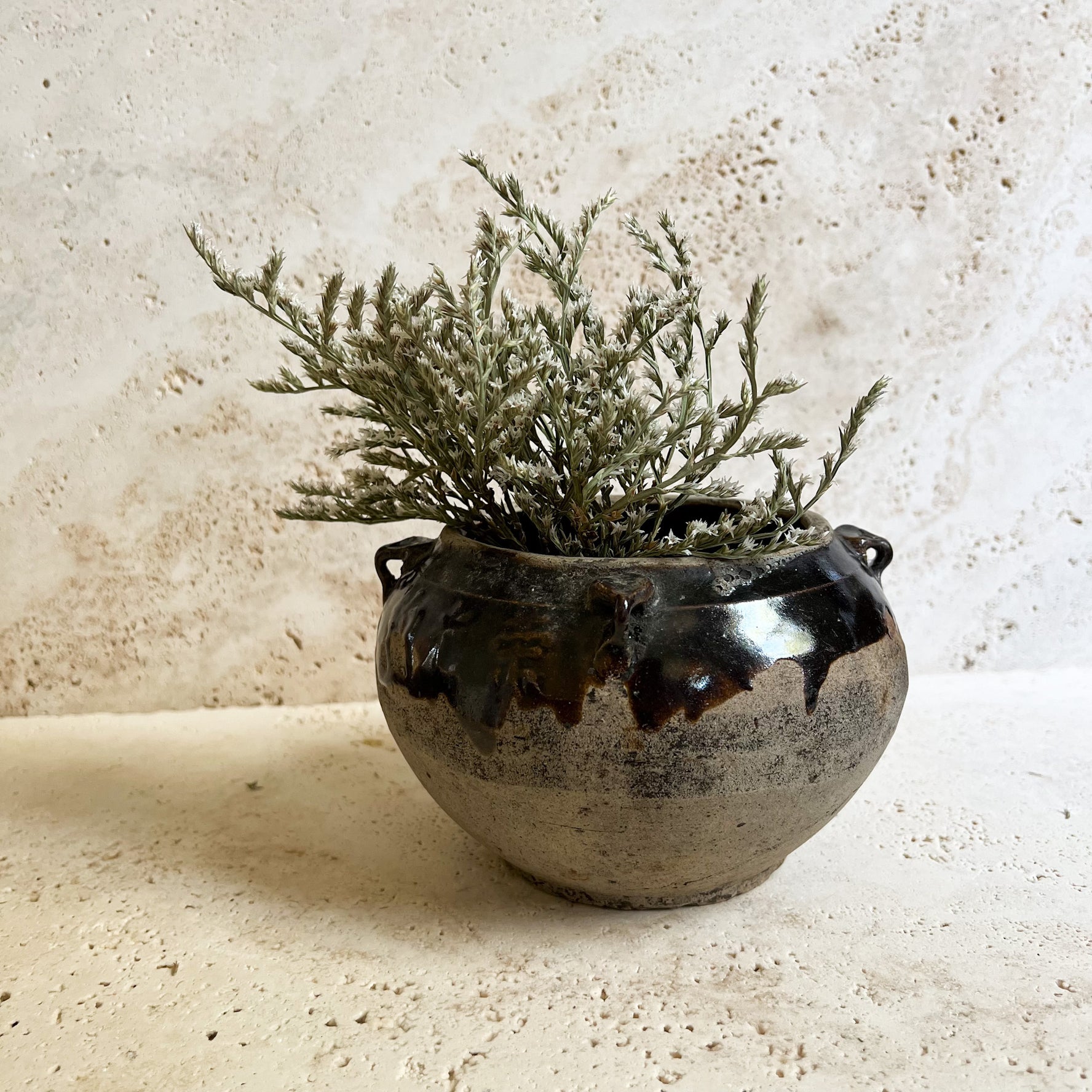 Antique brown Chinese pot filled with dried white German statice florals. The pot sits on a travertine stone.